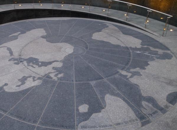 A map is laid out with various colored stone to create a floor and walkway. The coppery walls are lined with back-lit shapes that appear to coincide with various religious beliefs and institutions. 