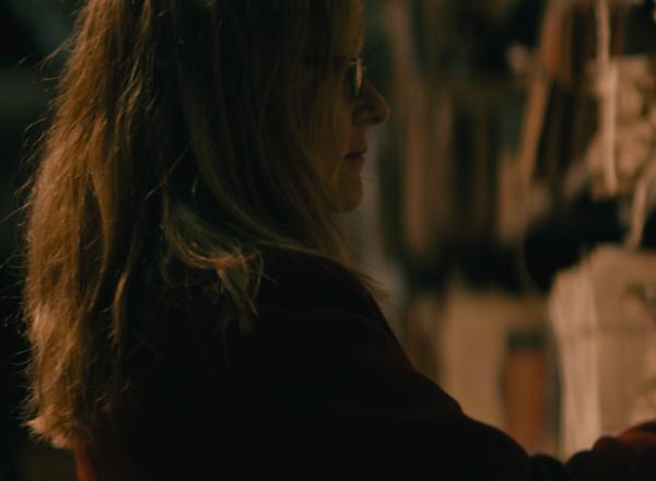 Lynda is seen in dark room, looking at shelf packed with documents. 